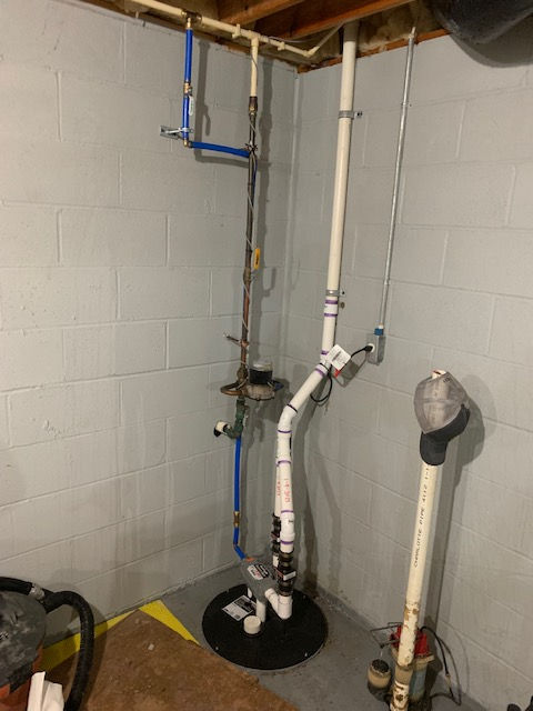 Sump pump system with white PVC discharge pipes and blue PEX water lines against a grey basement wall.
