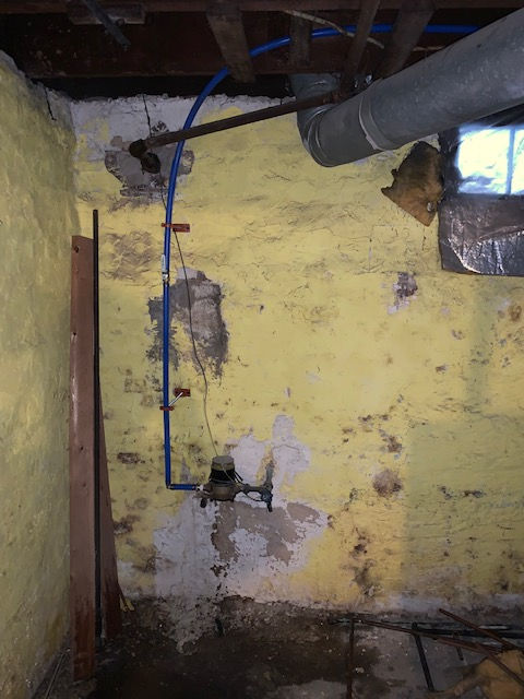 A blue water pipe and utility meter mounted on a yellow basement stone wall with exposed ceiling joists and ductwork.