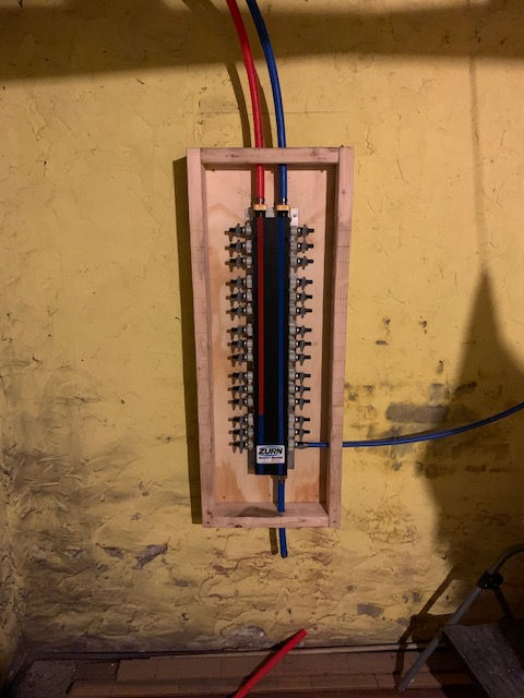 A plumbing manifold mounted in a wooden frame on a yellow wall, with red and blue PEX pipes connected at the top and bottom.
