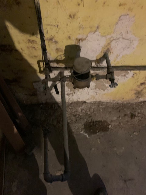 A residential water meter and pipe assembly mounted on a damaged, pale yellow interior basement wall.