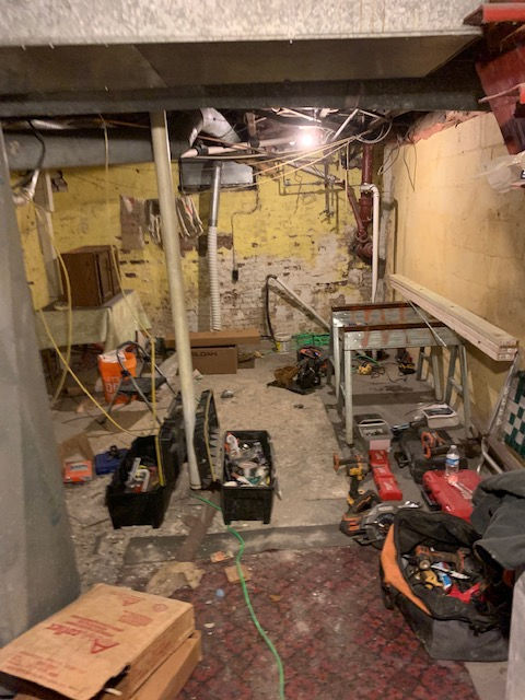 A cluttered, unfinished basement with yellow walls, exposed pipes, toolboxes, and construction materials on the floor.