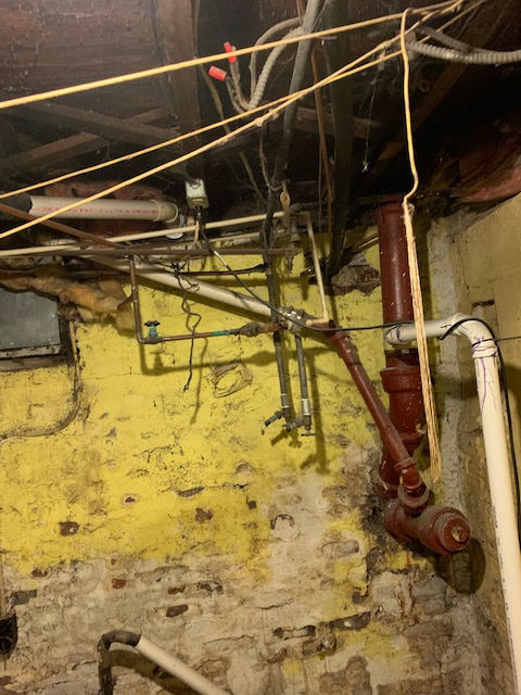 Exposed pipes, wiring, and a vertical drain stack against a yellow, textured foundation wall in a dimly lit basement.