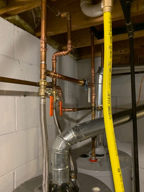 A basement utility area showing water heater plumbing, including copper pipes, orange-handled valves, and a yellow gas line.
