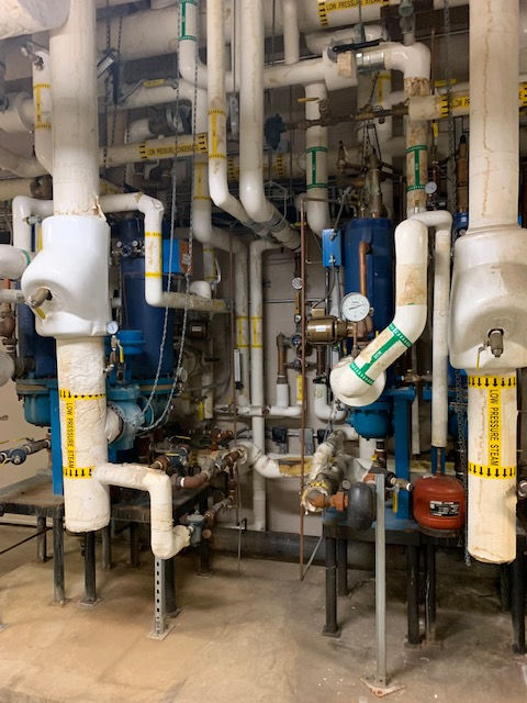 A mechanical room featuring large industrial pipes with white insulation and yellow labels connected to blue tanks.