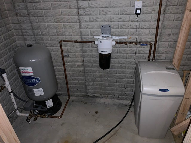 Water filtration system with a gray pressure tank, wall-mounted filter, and a water softener unit against a brick wall.