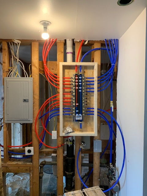A plumbing manifold with red and blue PEX tubing mounted on wooden studs next to an electrical panel in an open wall.