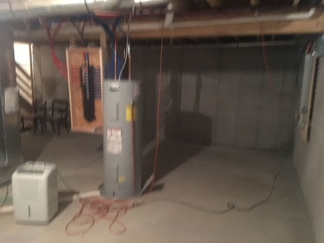 A basement with a grey water heater, a portable dehumidifier, an electrical panel on a wooden board, and concrete walls.