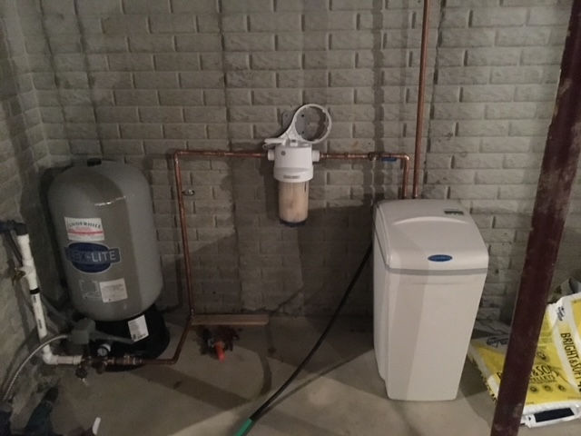 A grey pressure tank, a central water filter, and a white water softener unit installed against a grey brick wall.