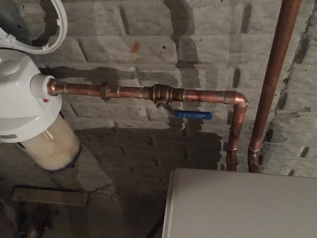 A water filtration unit with a blue handle shut-off valve connected by copper pipes against a concrete block wall.