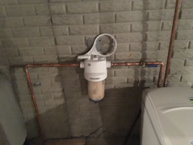 A water filter housing is mounted on a brick wall between copper pipes, with a hanging wrench tool above it.