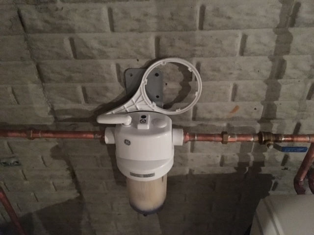 A white water filter housing installed on a brick wall, connected to copper pipes with a detached mounting bracket above.