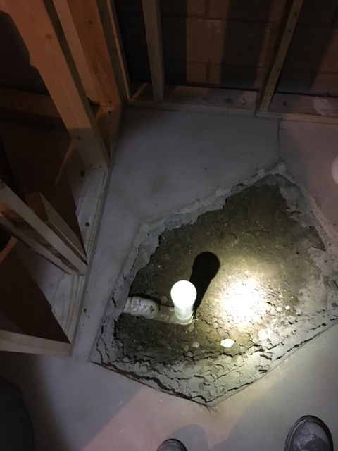 A hole cut in a concrete floor reveals a vertical white plumbing pipe inside a wooden-framed room under construction.