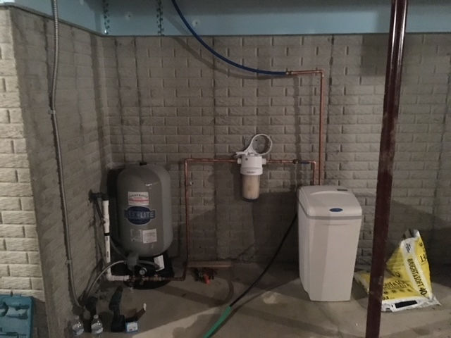 A residential water filtration system with a gray pressure tank, a wall-mounted filter, and a white water softener unit.
