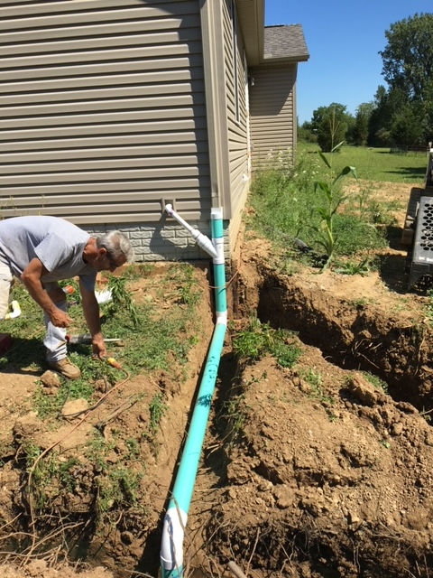 A person works on installing a light-blue PVC drain pipe connected to a house downspout inside an excavated trench.