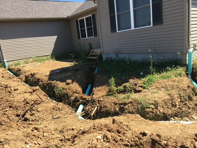 A house exterior featuring a freshly dug trench with blue PVC drainage pipes installed along the foundation.