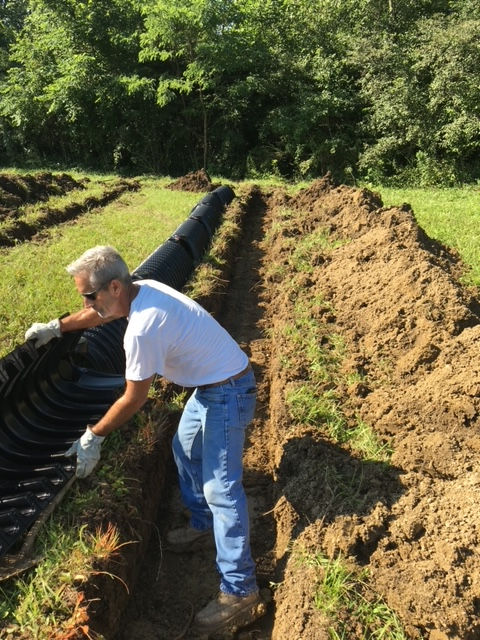 A person wearing work gloves and a white shirt places sections of black corrugated drainage pipe into a dirt trench.