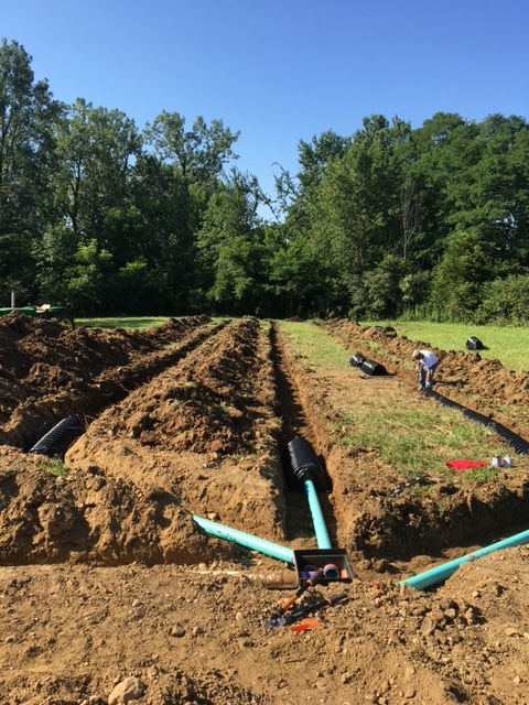 A worker installs pipes in trenches across a field, with piles of brown soil and green trees in the background.