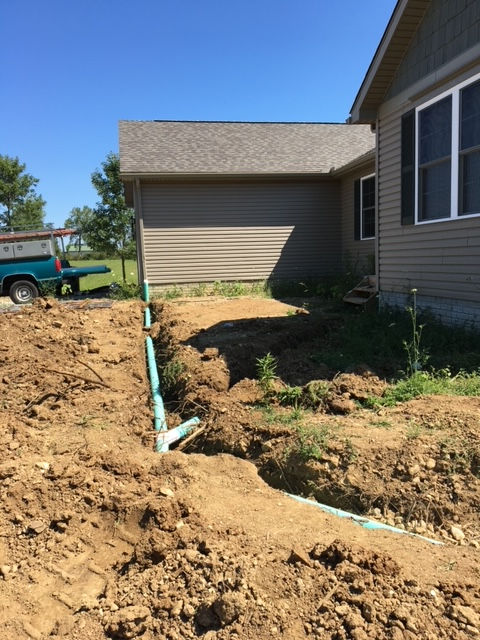 A trench with green drainage pipes runs alongside a house with brown siding and a driveway.