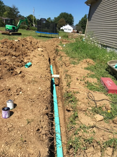 A construction site shows a green pipe installed in a deep trench running alongside the side of a residential house.