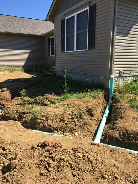 A trench containing a green drainage pipe running along the foundation of a tan, vinyl-sided house.