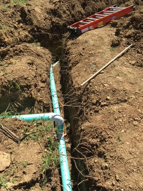 A blue PVC pipe is connected to a T-joint inside a dirt trench, with an orange ladder resting on the ground above.