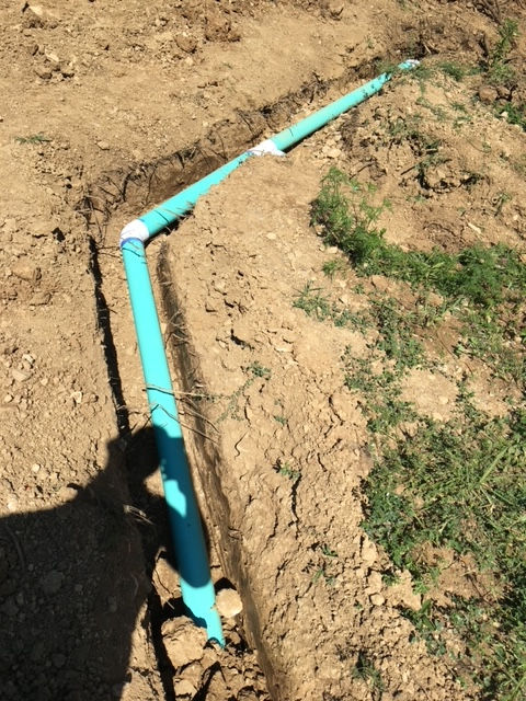 Light blue PVC piping is laid in a dirt trench, connected by a white elbow joint, viewed from above.