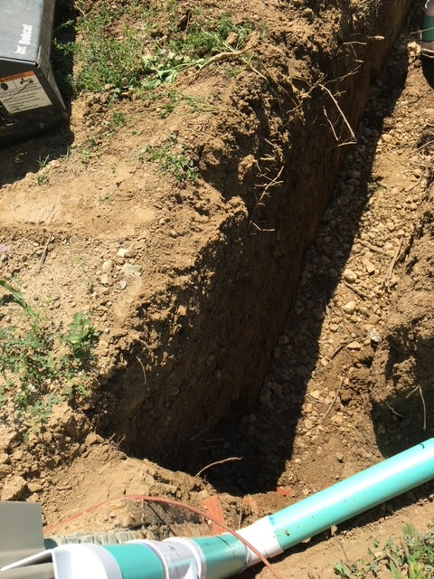 A turquoise PVC pipe sits at the bottom of a deep, narrow dirt trench excavated in a grassy area.