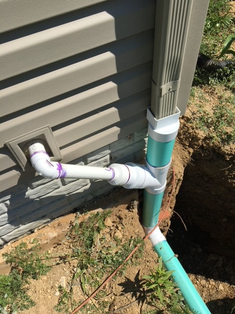 A white PVC pipe connects a house wall to a green downspout drain buried in the soil.