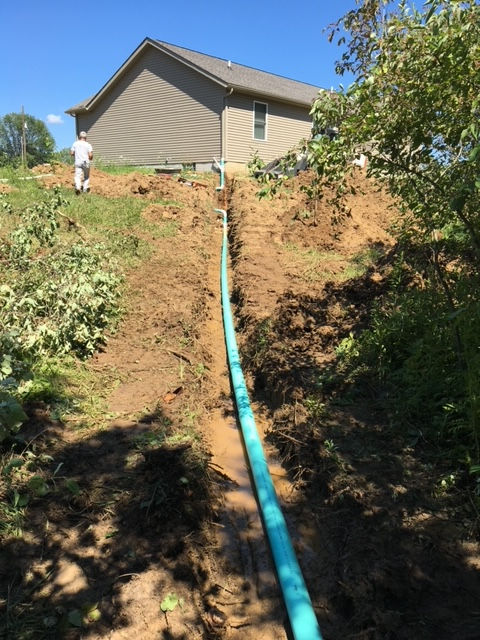 A long, light-blue pipe lies in a trench extending toward the side of a tan house on a sunny day.