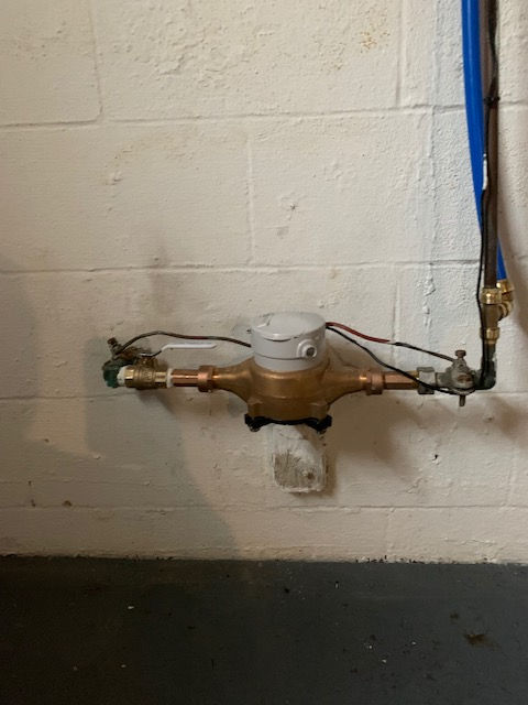 A residential water meter connected to copper pipes mounted on a white concrete block wall.