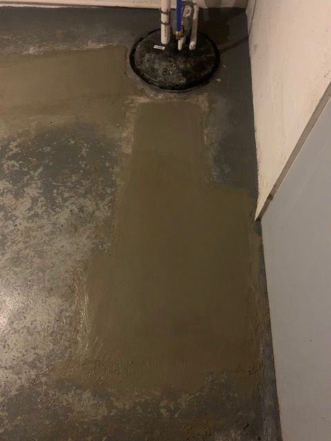 A patch of fresh, wet concrete on a grey garage floor surrounding a sump pump basin in the corner of a room.