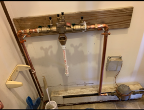 Copper plumbing pipes and a backflow preventer assembly mounted on a wooden board against a wall near a water meter.