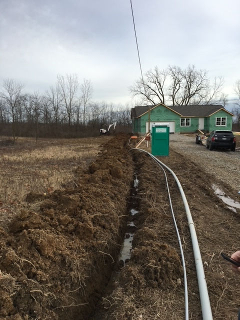 A long trench dug into a field leads toward a house under construction, with pipes lying next to the open ground.