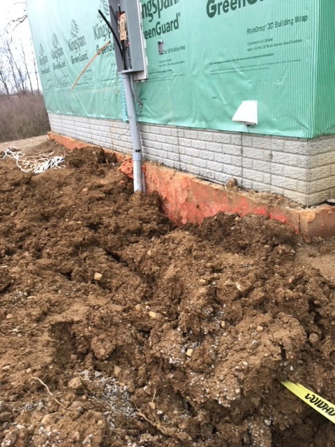 A utility meter mounted on the exterior of a building under construction, with a trench dug in the dirt alongside it.