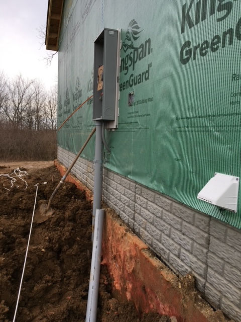 An electrical meter box installed on the exterior of a construction site with green sheathing and brick foundation.
