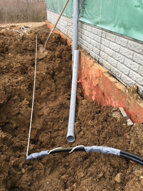 An outdoor construction site shows a grey PVC pipe against a brick foundation, with electrical cables lying in the dirt.