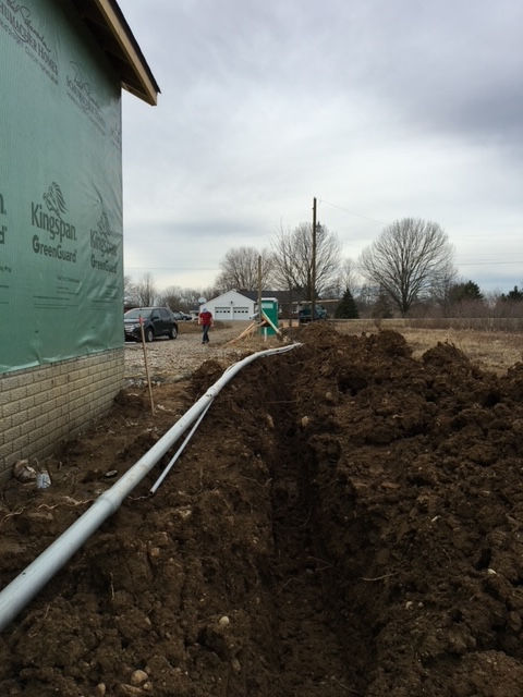 A grey pipe runs along an excavated trench next to a house with green wall sheathing under a cloudy sky.