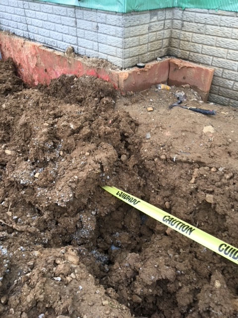 A construction site trench runs alongside a building foundation with a yellow caution tape stretched across the opening.