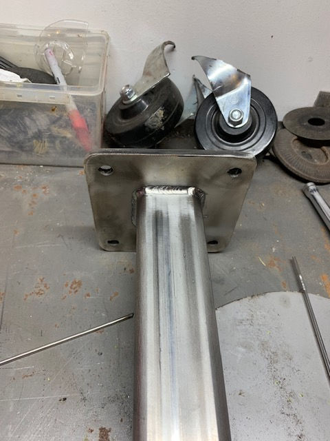 A square metal mounting plate welded to a rectangular steel tube, with two caster wheels resting in the background.