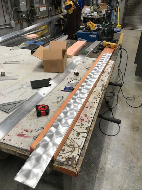 A metal strip with an engine-turned finish rests on a wooden block atop a workshop table with tools and supplies nearby.