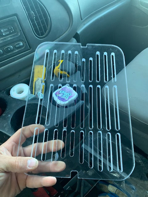 A person holds a clear, rectangular acrylic refrigerator shelf or vegetable crisper cover with ventilation slots.