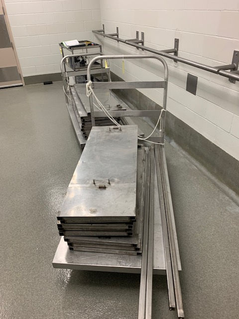 Metal carts holding stacked rectangular metal plates and long metal rods in a sterile room with gray flooring and white walls.