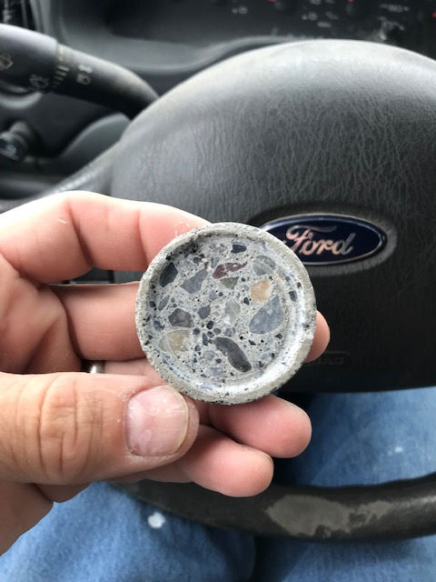 A person holding a small, circular piece of concrete with visible aggregate, set against a Ford steering wheel background.