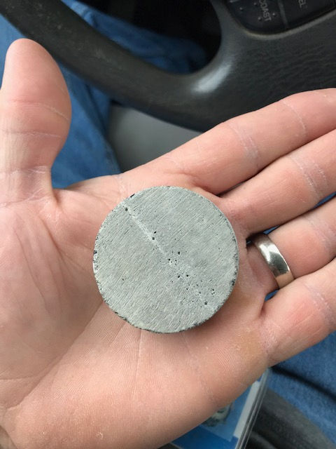 A hand holds a small, circular, gray concrete disc featuring a thin crack and small surface pores.