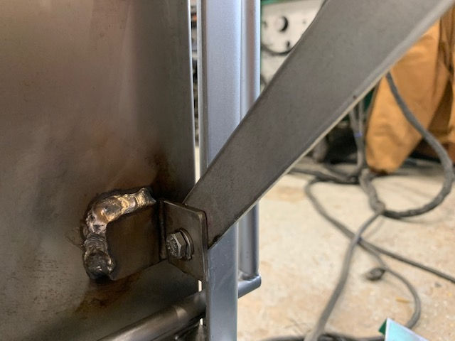 A metal bracket welded to a stainless steel panel, with a flat metal brace attached by a bolt and nut.