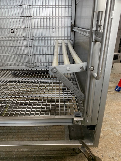 A close-up of a metal cage interior featuring a three-rod folding shelf attached to the side wall above a grid floor.