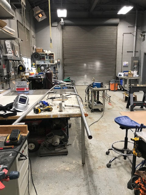 A well-lit fabrication workshop featuring metal poles on a workbench, welding equipment, tools, and a large bay door.