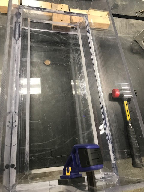 Clear acrylic rectangular container being assembled with a blue clamp and a rubber mallet on a dark workbench.