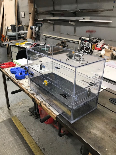 A clear acrylic box sits on a workbench in a workshop, containing a dark, textured base and a yellow object.