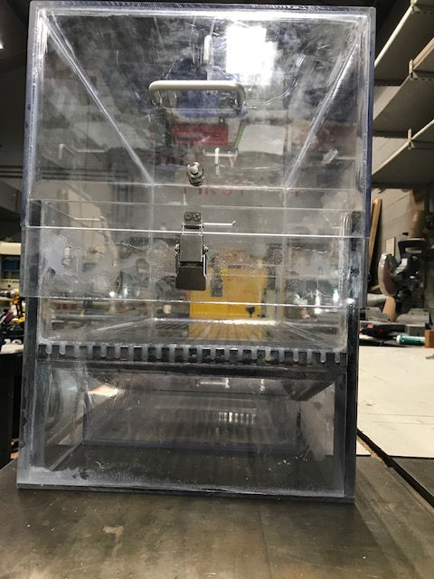 A clear acrylic display case with a hinged front lid and a slotted bottom shelf, sitting on a workbench.